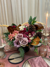 Load image into Gallery viewer, A floral centrepiece with a mix of red, pink, and white flowers, green foliage, and pine cones, arranged in a round, dark vase, displayed on a table with a purple tablecloth.