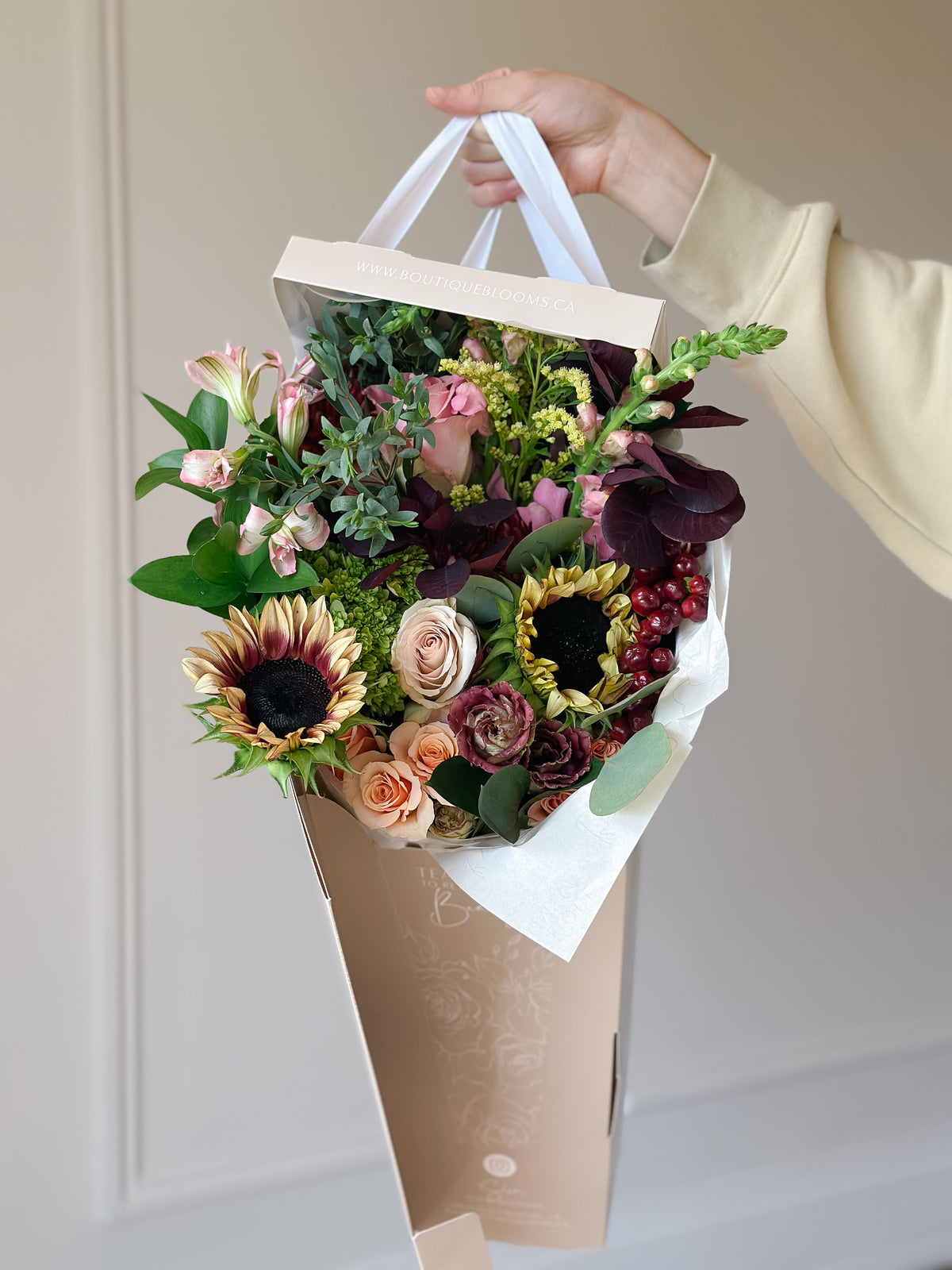 A hand holding a large floral arrangement in a kraft paper bag. The bouquet features sunflowers, pink and burgundy roses, green foliage, and small berry accents, with white paper handles visible at the top against a neutral indoor background.