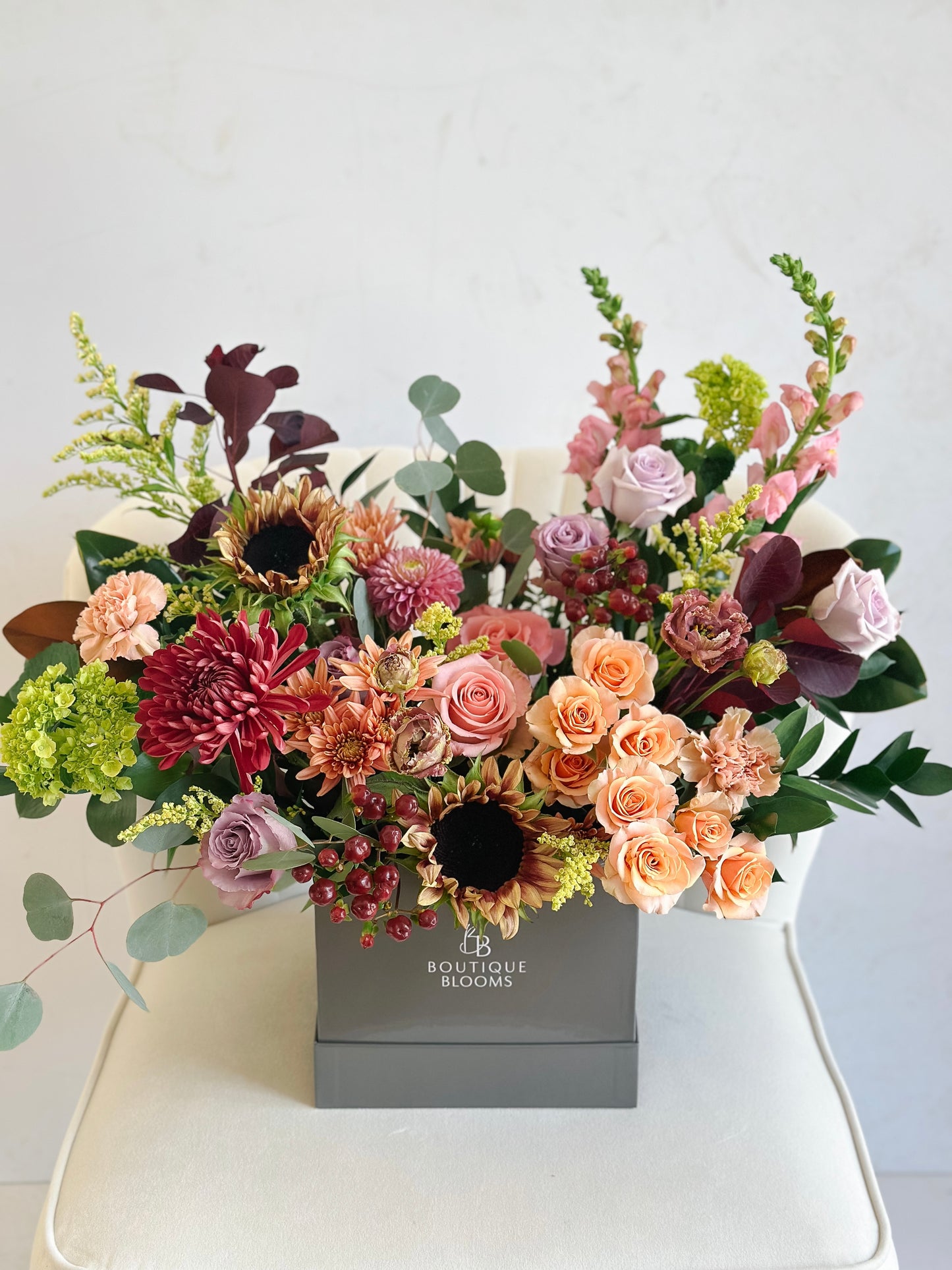 A vibrant autumnal floral arrangement in a square gray box from Bougie Blooms, featuring peach roses, burgundy chrysanthemums, sunflowers, and green foliage on a white chair.