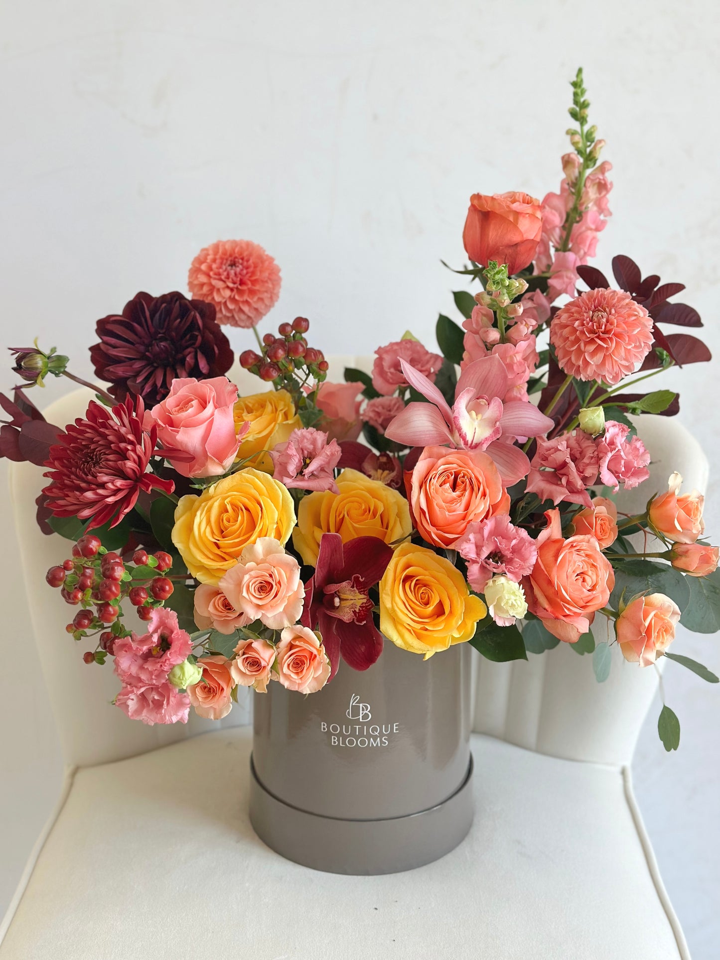 A vibrant floral arrangement featuring yellow, pink, orange, and burgundy flowers in a round gray 'Boutique Blooms' box, set on a white chair against a white wall.