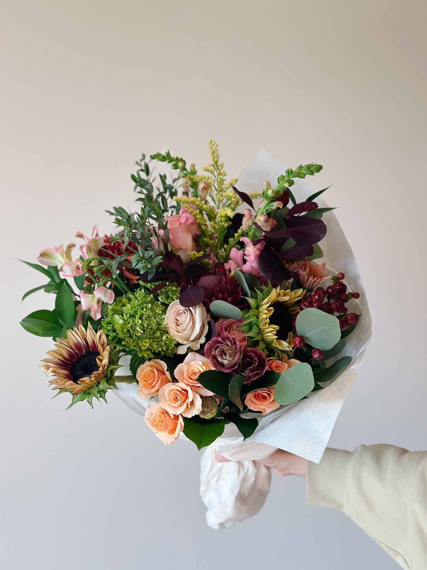 A hand holding a large, beautifully arranged floral bouquet featuring sunflowers, pink and peach roses, burgundy dahlias, and green foliage, wrapped in white paper against a plain beige background.