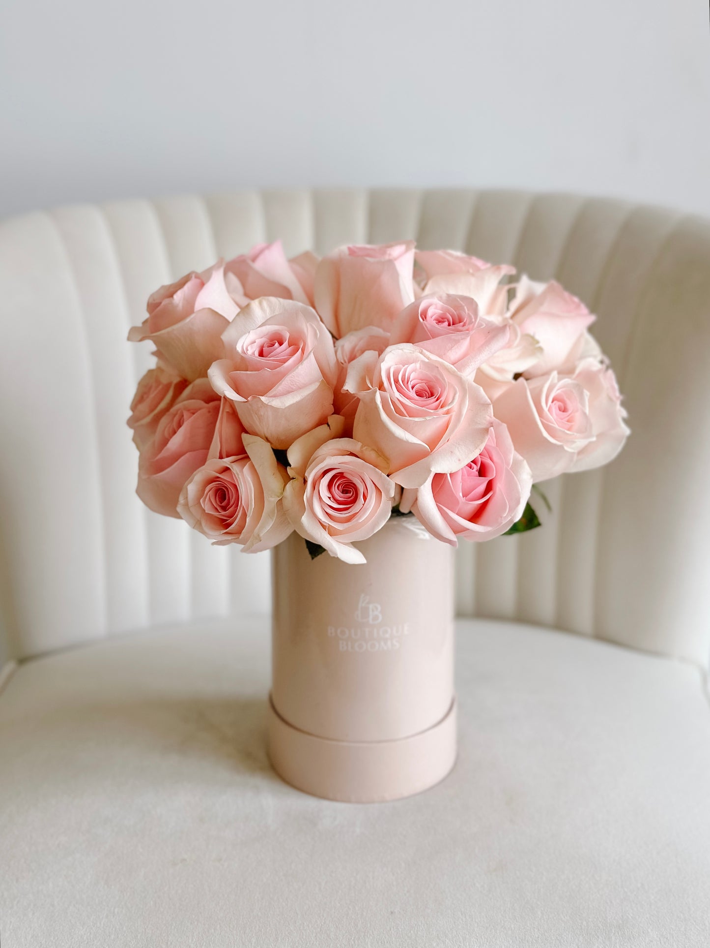 Bouquet of pink roses in a cylindrical pink container on a white surface with a white chair in the background.