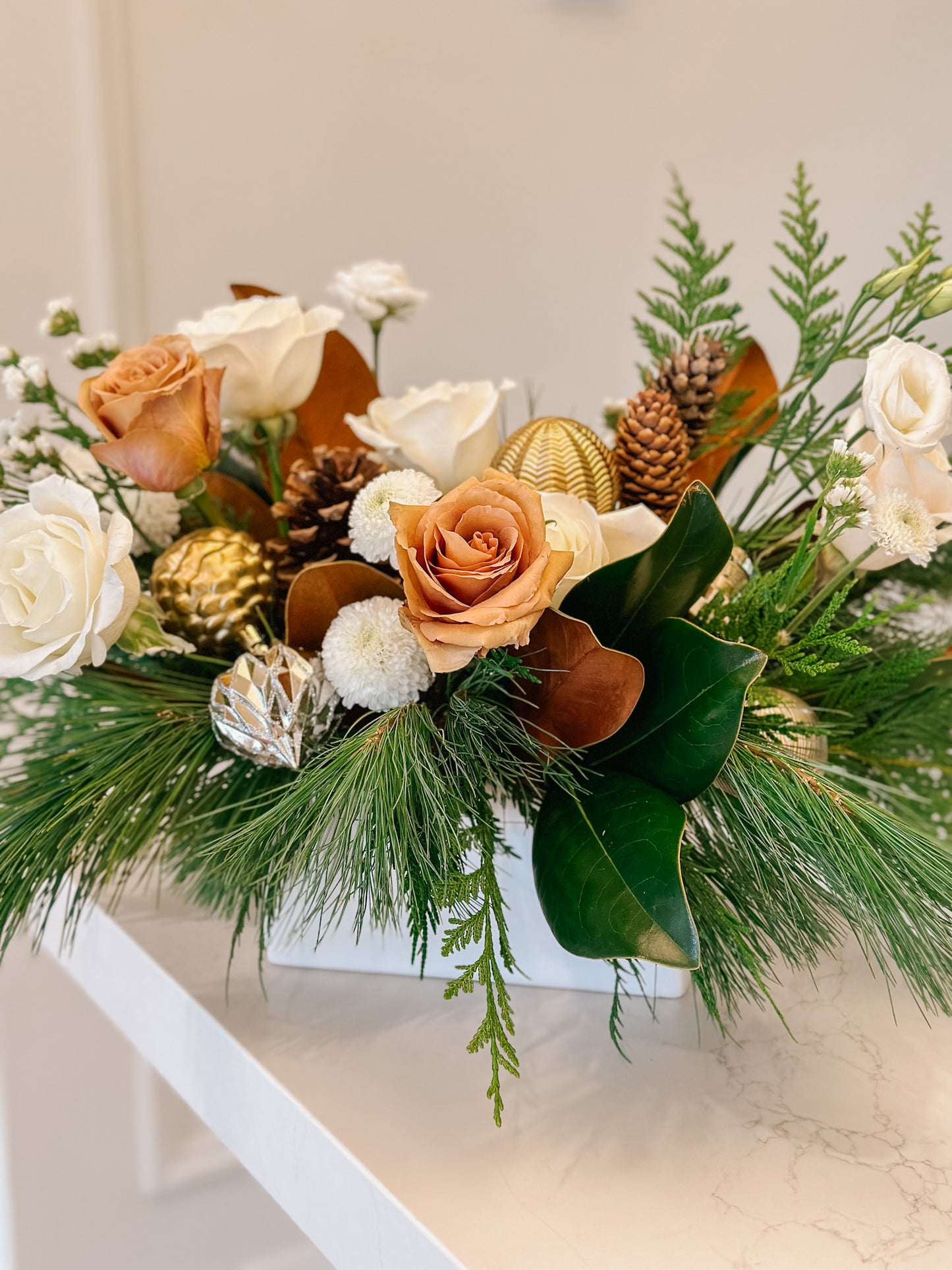 a beautiful winter centerpiece in a white ceramic pot featuring white roses, toffee roses, winter greens, and a variety of christmas ornaments.