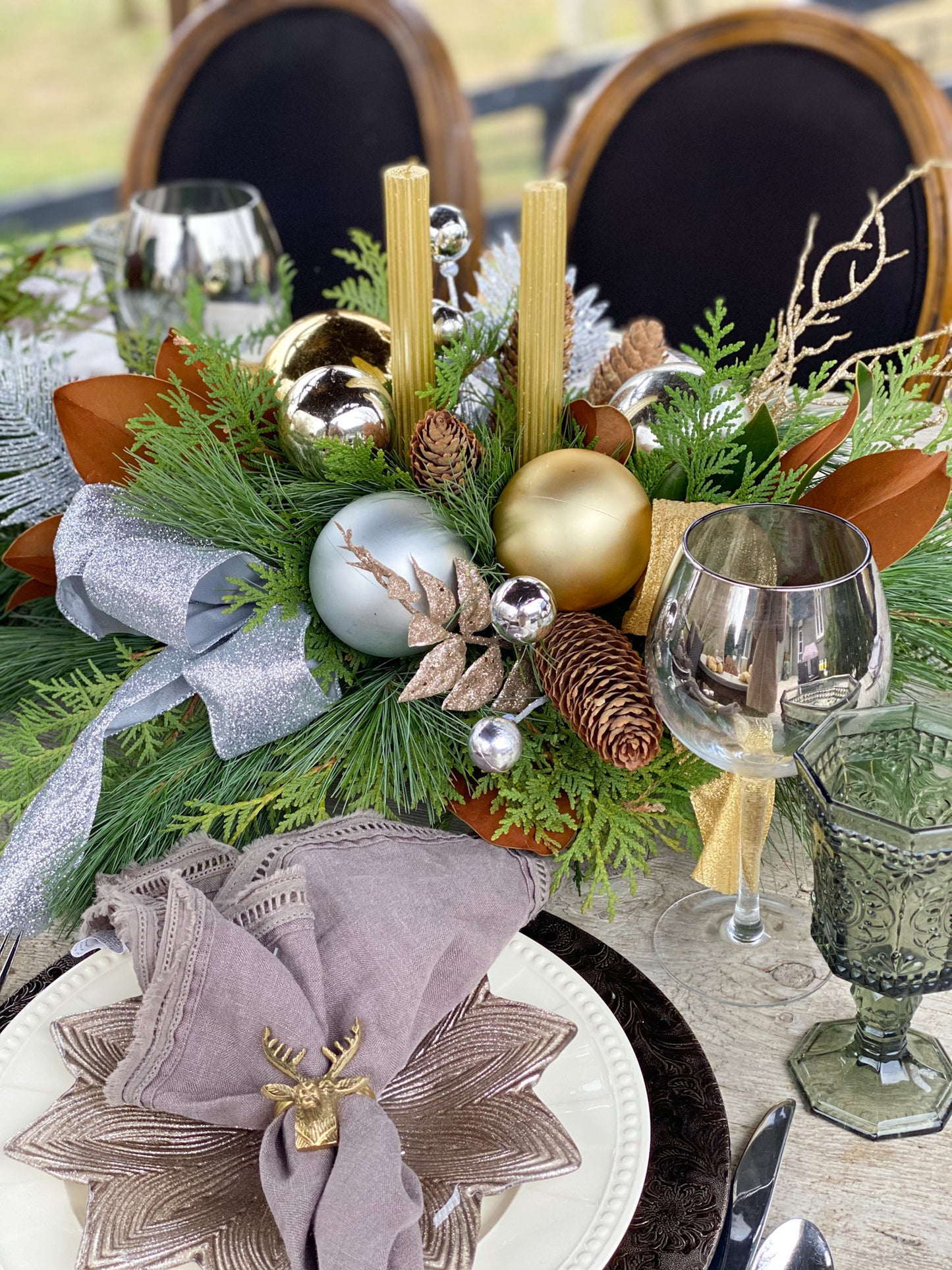 A holiday table centerpiece featuring gold and silver ornaments, pine, cedar and magnolia foliage, pinecones, and golden candle sticks, arranged in a bed of fragrant pine needles.