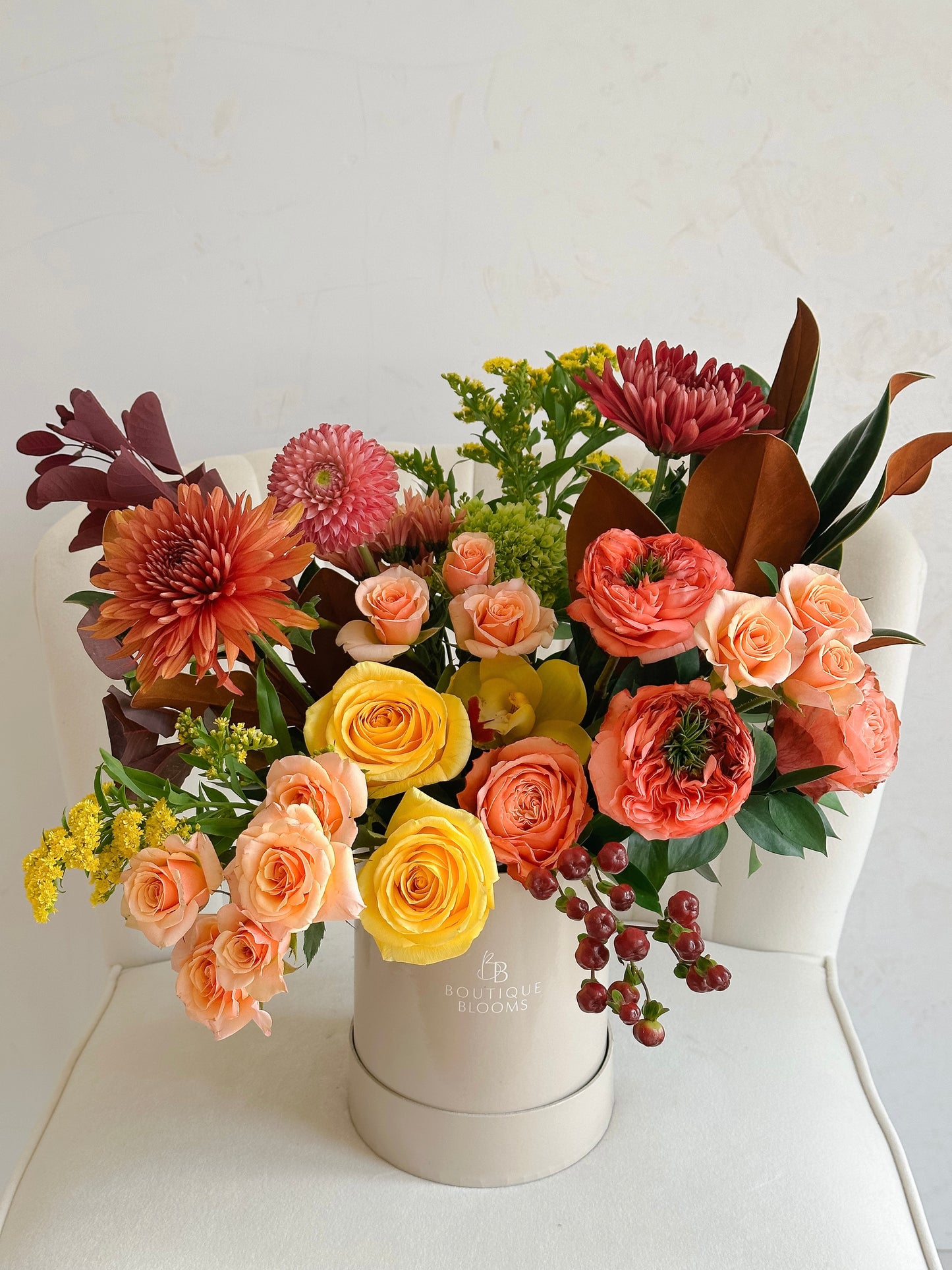 A vibrant autumn floral arrangement in a round white box, featuring yellow and peach roses, orange chrysanthemums, red ranunculus, and green foliage, displayed on a white chair against a light wall.