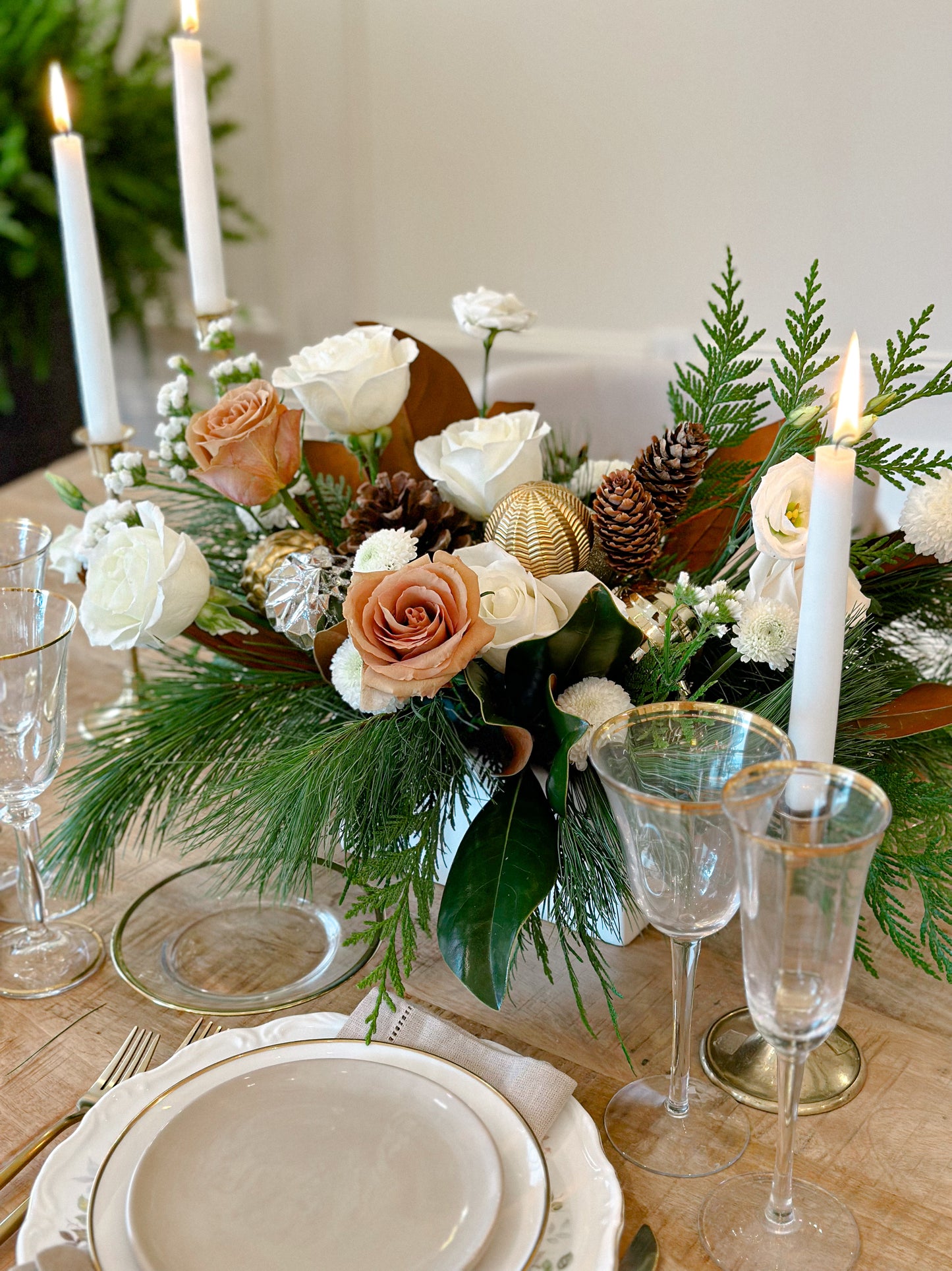A centerpiece featuring a clear vase with a floral arrangement that includes white and pink roses, green pine needles, and other decorative elements, placed on a table with a white plate and candles in the background.