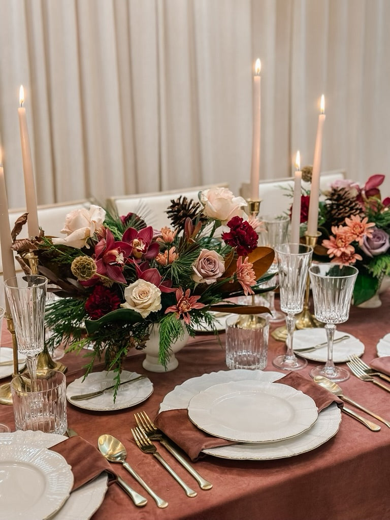 A holiday centerpiece featuring a mix of red and pink flowers and green foliage, placed on a table with white plates, silver cutlery, and lit candles.