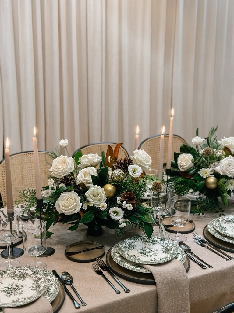 A festive floral centerpiece with creamy white blooms and seasonal foliage, arranged on a table with plates and silverware, suitable for holiday celebrations.
