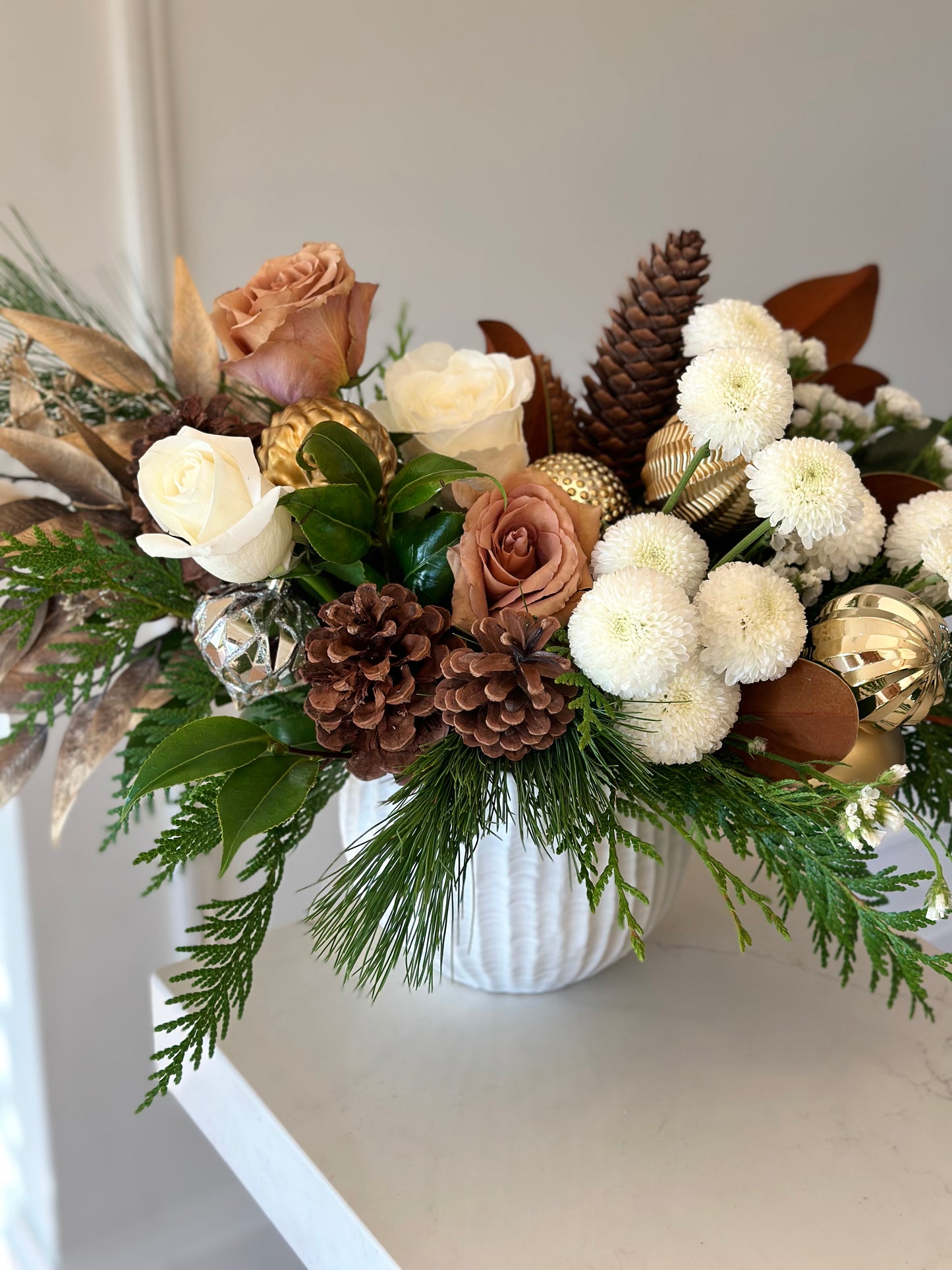 A decorative bouquet of flowers featuring a variety of holiday-themed flowers in a white vase, including gold-toned ornaments and greenery.