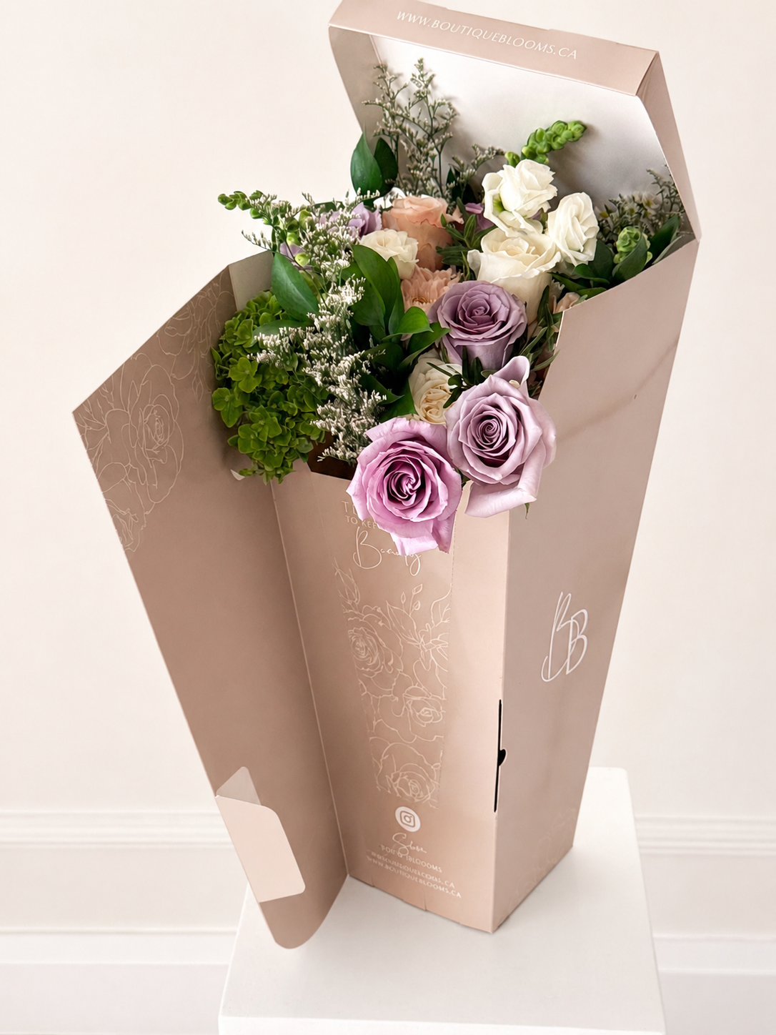 Lavender roses, ivory blooms, and green hydrangea arranged in a luxury Boutique Blooms flower bag with fresh foliage.