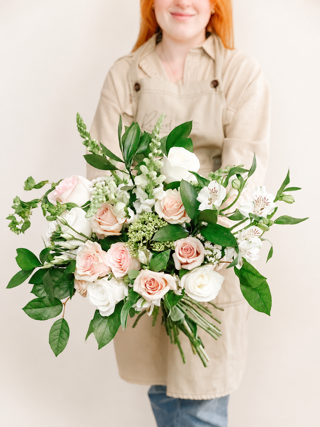 Cream and blush roses with white seasonal blooms arranged in a modern floral gift bag with lush greenery.