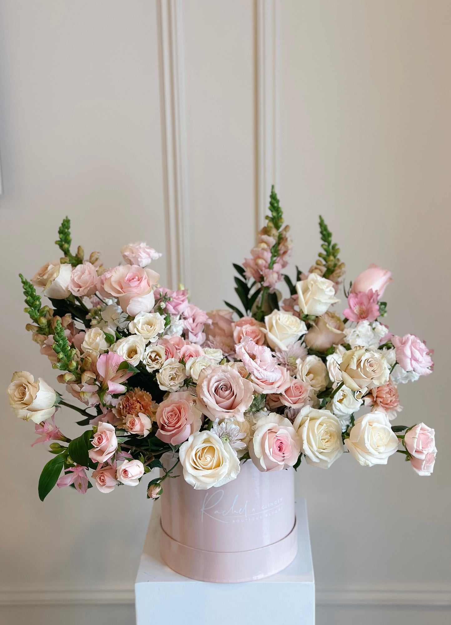 A round hat box arrangement filled with an assortment of blush and pink flowers with hints of foliage.