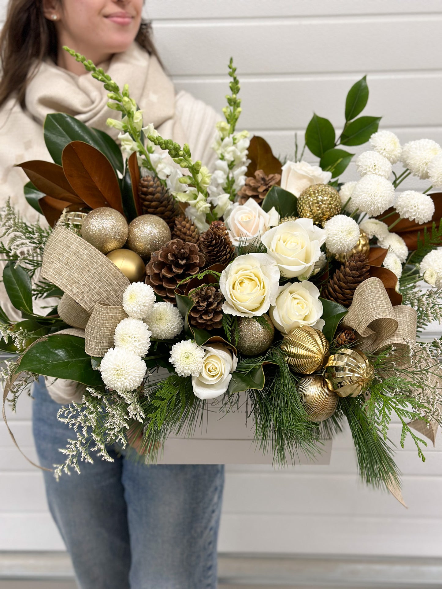A person holding a large floral arrangement featuring white and cream flowers, green foliage, brown pinecones, and decorative elements in a gold container.