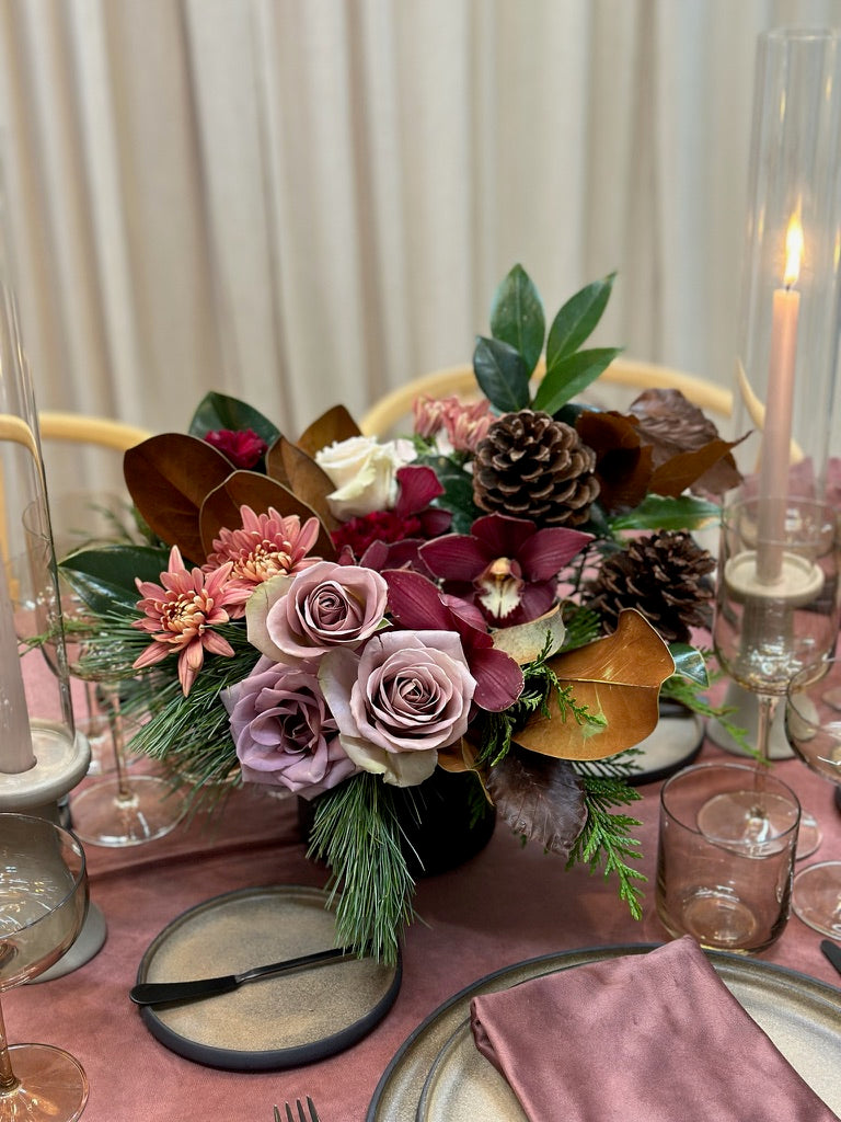 A floral centrepiece with a mix of red, pink, and white flowers, green foliage, and pine cones, arranged in a round, dark vase, displayed on a table with a purple tablecloth.