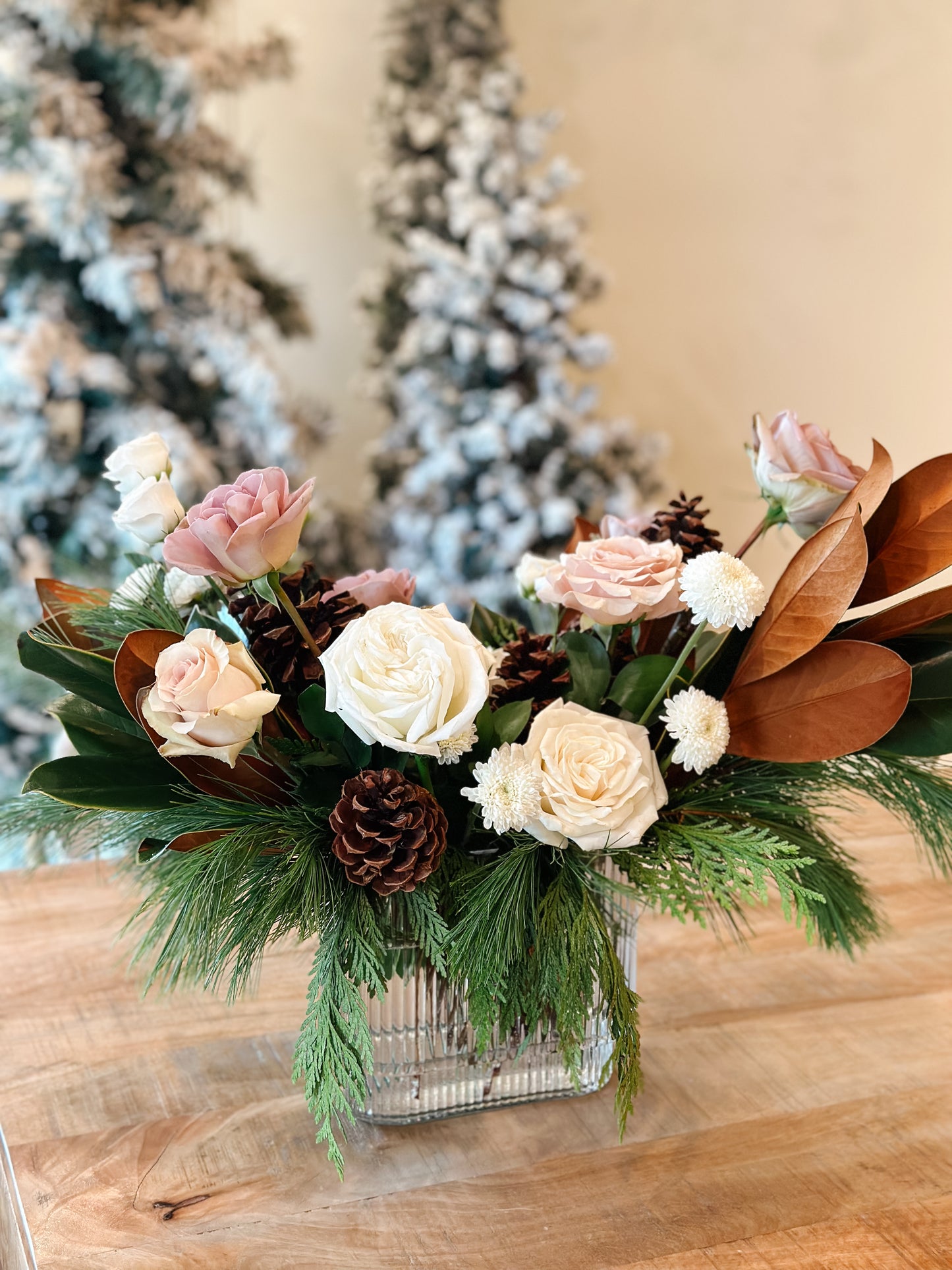 Floral arrangement with roses and pinecones in a clear vase on a wooden surface, with a blurred Christmas tree in the background.