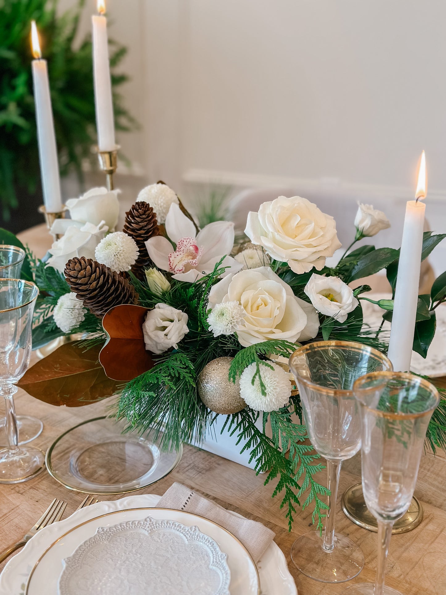 A centrepiece featuring a white orchid, white roses, and greenery, with pinecones and ornaments, displayed on a table with lit candles and a festive tableware setup in the background.