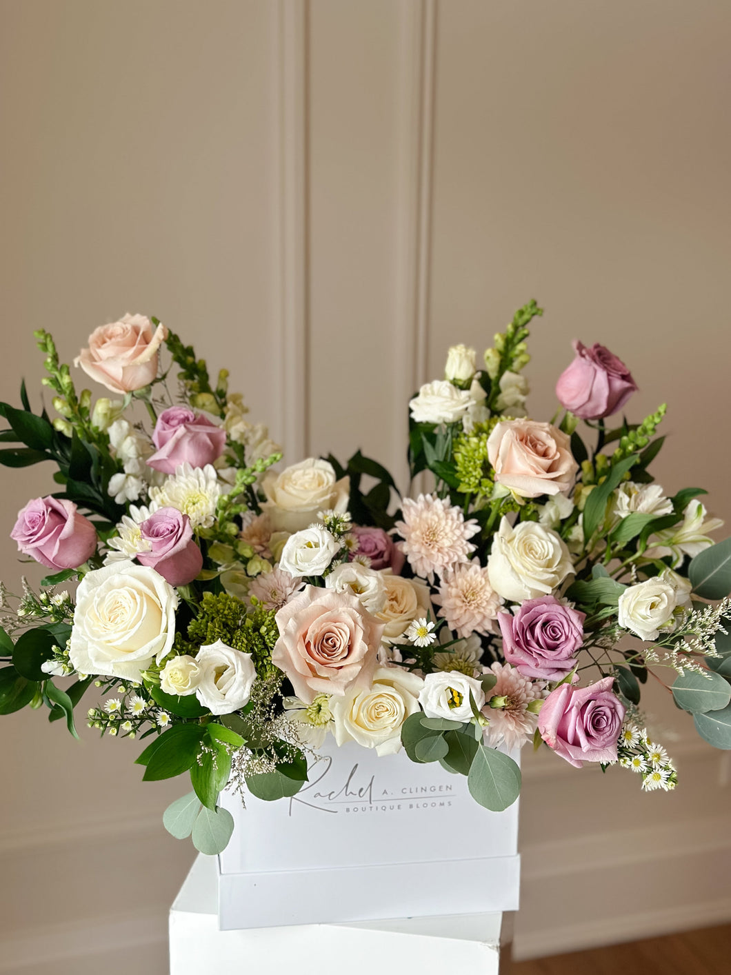 A medium-stemmed floral arrangement featuring pastel mauves, soft pinks, and fresh greenery in a bloom box.