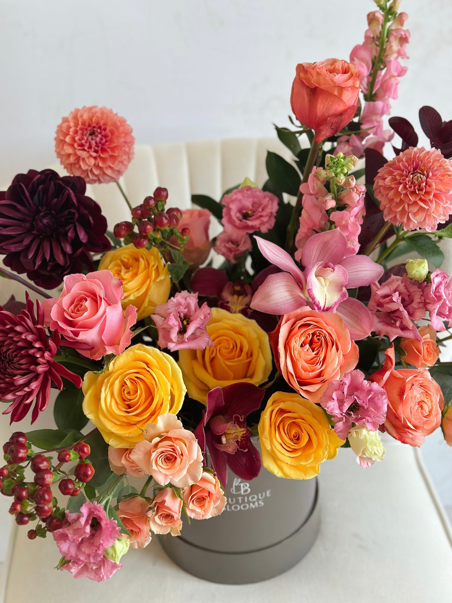 A vibrant, multicolored floral arrangement featuring yellow and orange roses, pink carnations, orchids, and dahlias, presented in a round gray gift box with 'BOUTIQUE BLOOMS' branding, set against a neutral white background.