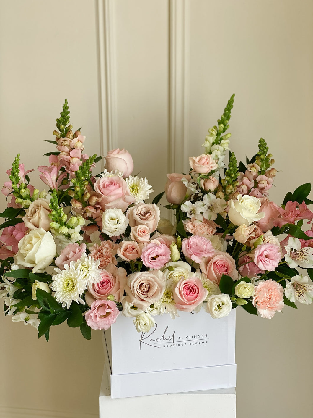 A large, elegant floral arrangement featuring a variety of premium blooms in shades of pink, white, and green, presented in a white box with 'Rachel' branding on the side.