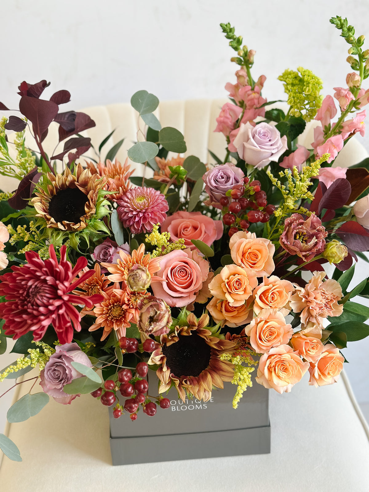 A vibrant, multicolor floral arrangement featuring sunflowers, roses, chrysanthemums, and green foliage, presented in a gray square box with the text 'OUT OF THE BLOOMS' visible on the front.