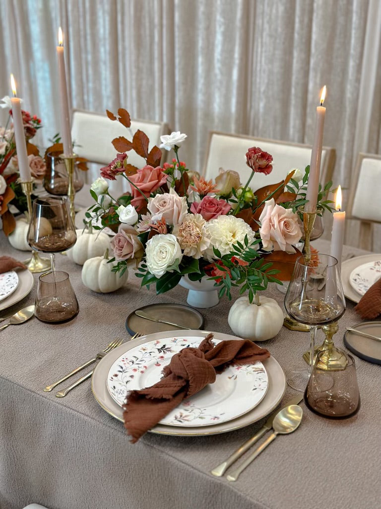 A thanksgiving table setting featuring an autumn centrepiece with floral in a vase, surrounded by white pumpkins, with plates, cutlery, and candles on a tablecloth.