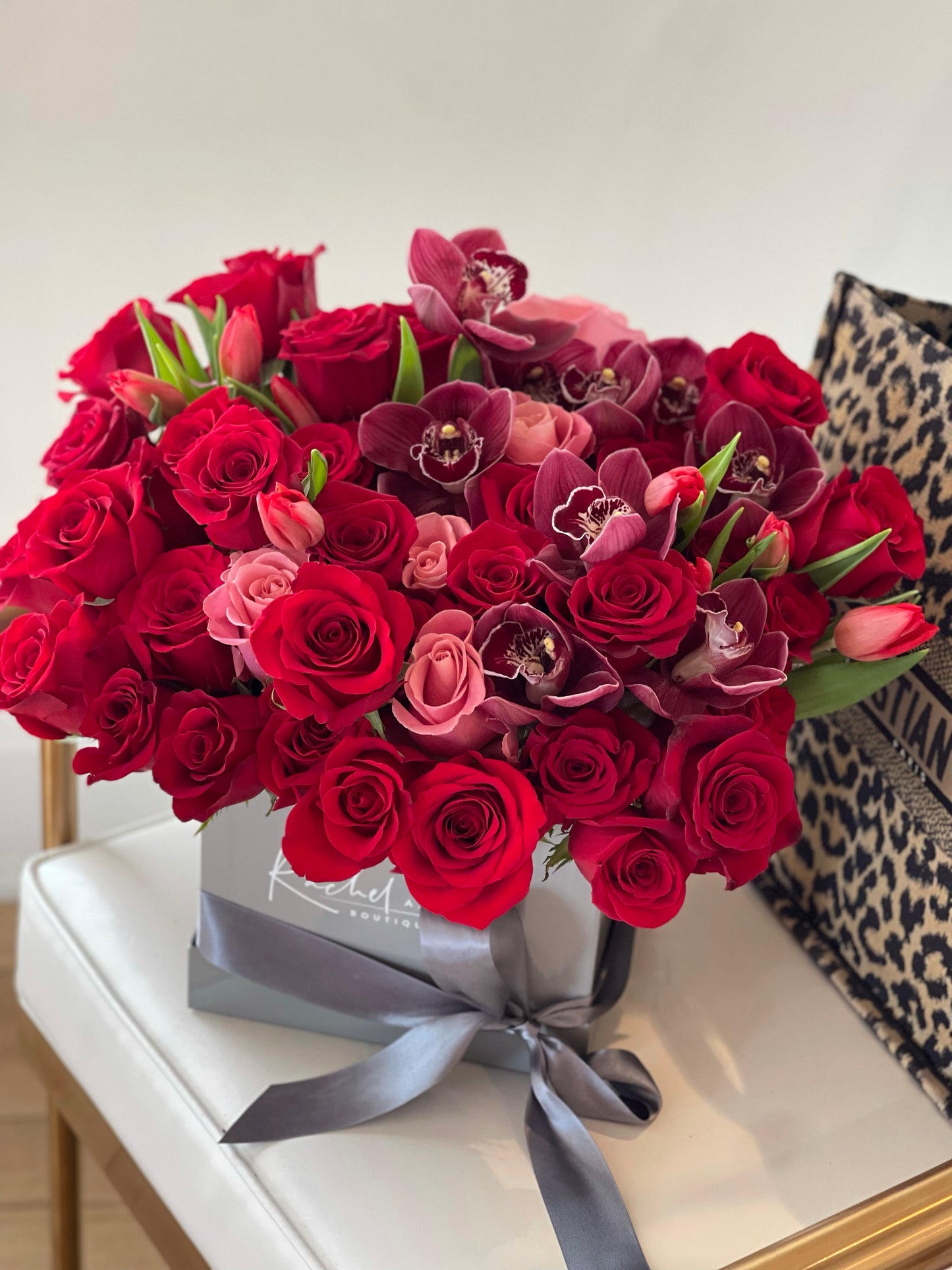 A bouquet of red roses, tulips, burgundy orchids, and pink roses arranged in a clear box with a grey ribbon.