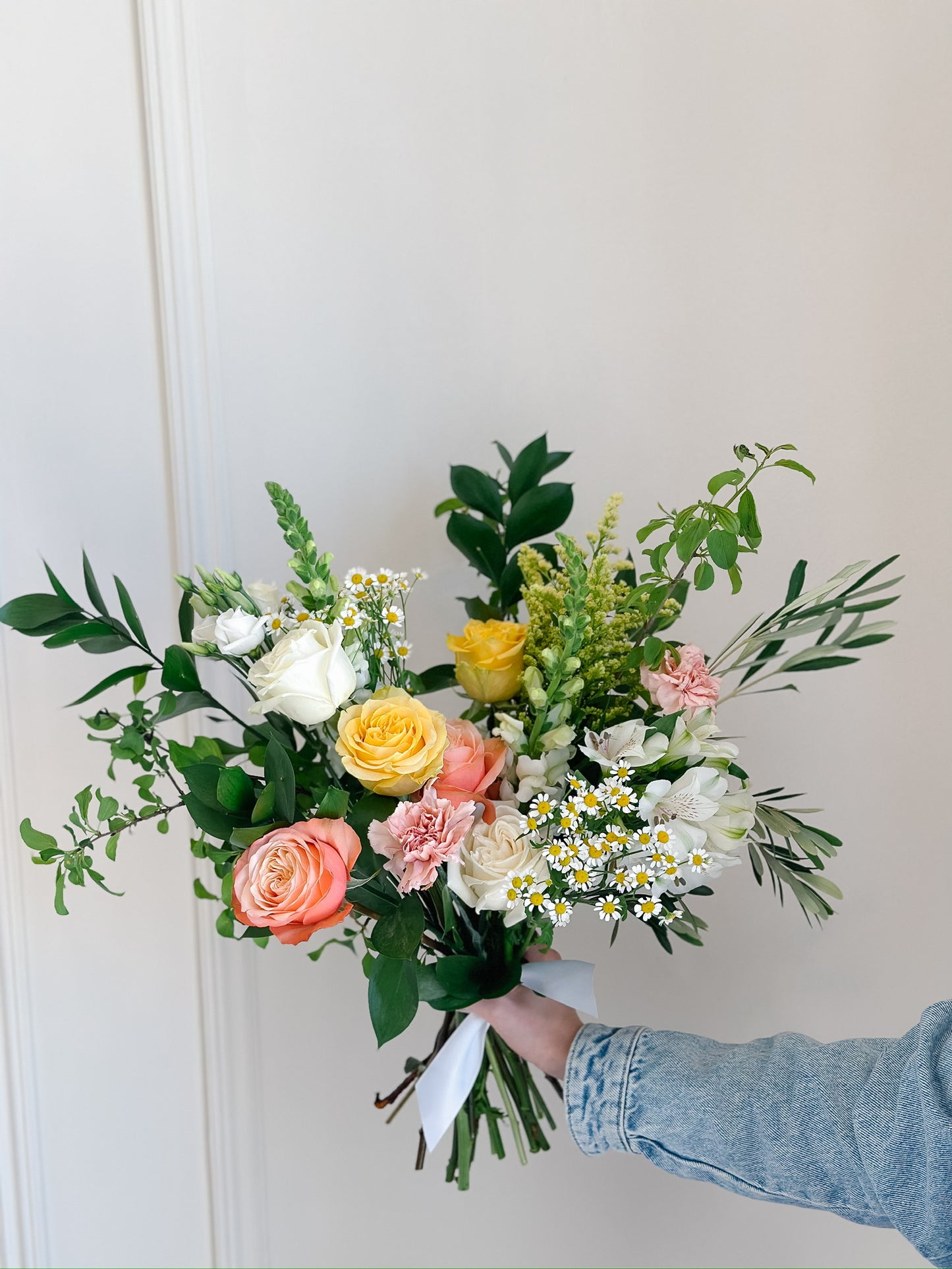 Hand-tied bouquet featuring peach and yellow roses, white chamomile blooms, and lush greenery tied with a satin ribbon.