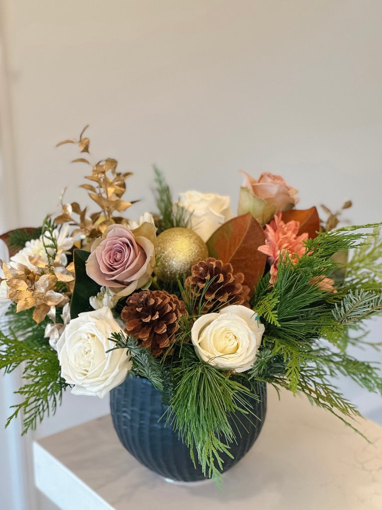 A floral arrangement featuring roses, pinecones, and greenery in a blue vase, with a neutral background.