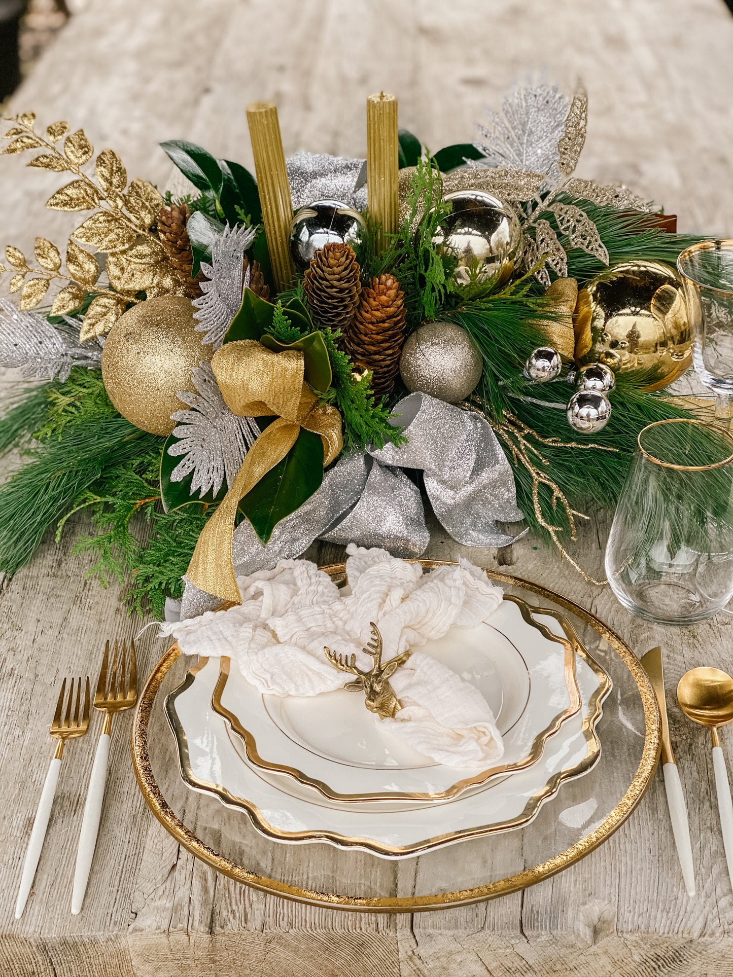 A festive holiday table centerpiece with a mix of gold and silver ornaments, pine, cedar and magnolia, pinecones and gold candlesticks, arranged on a bed of white tissue paper on a plate.