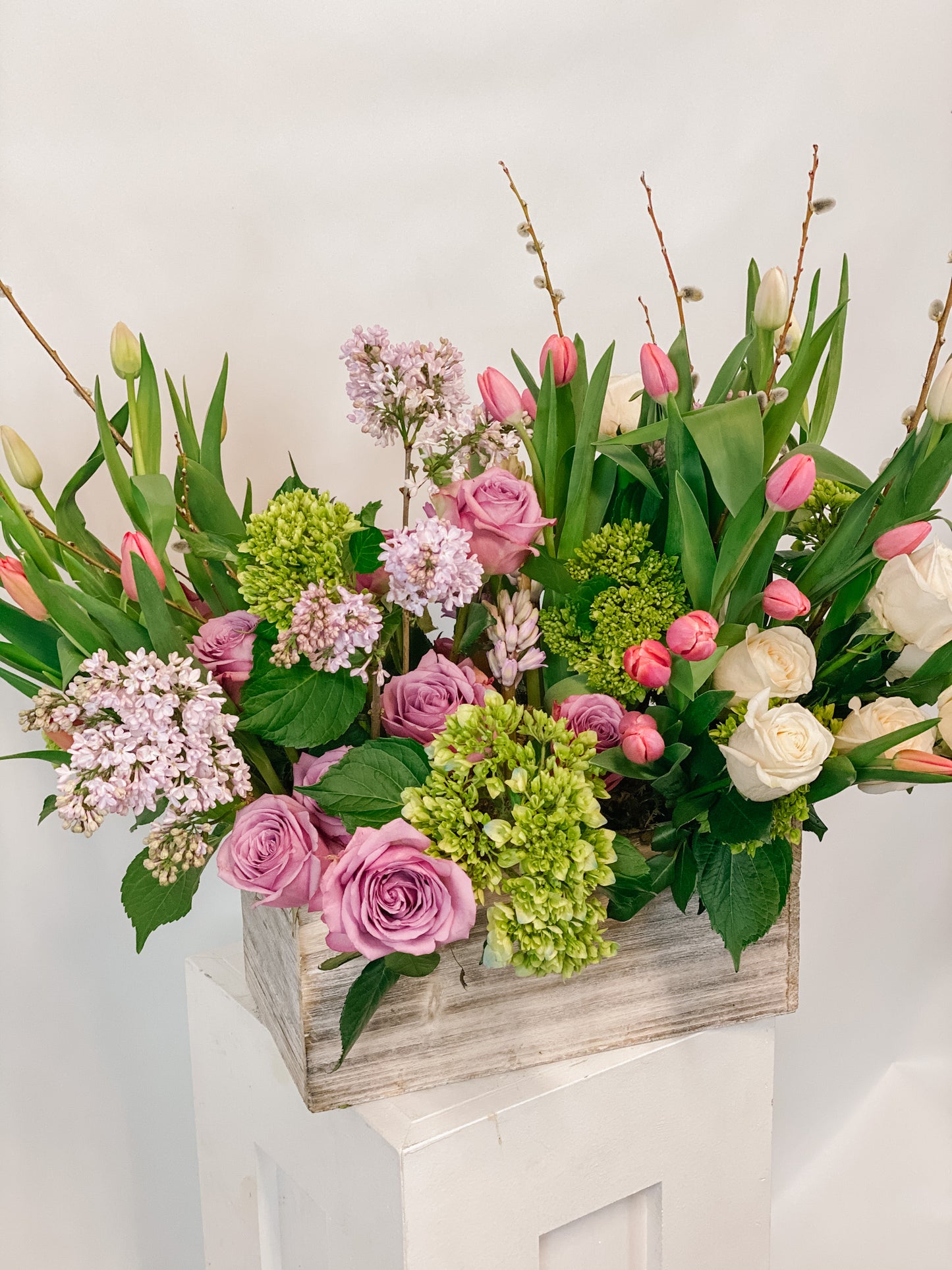 A wooden box arrangement of various flowers including roses, hydrangea, and tulips, with a color combination of pink, green, and white.