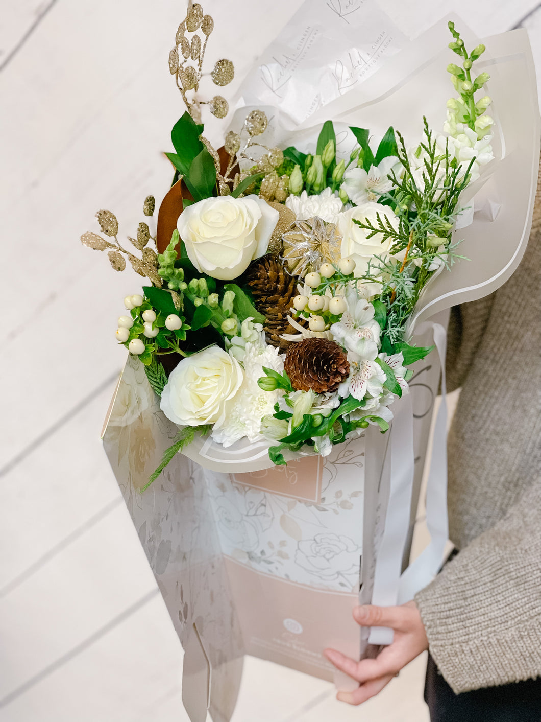 A hand-tied bouquet of white florals including roses and pinecones, wrapped in tissue paper with decorative elements.