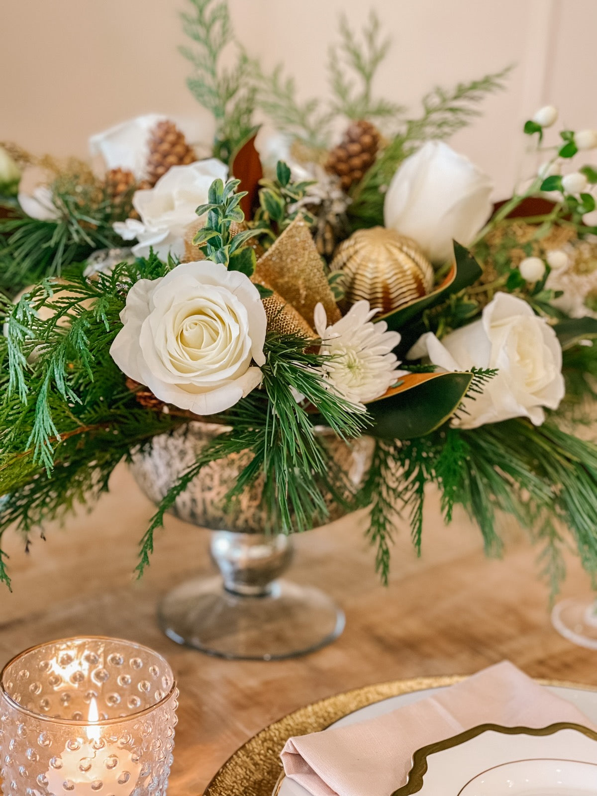 a festive holiday centerpiece with white flowers, fresh winter greens, gold ornaments and ribbon, and pinecones.