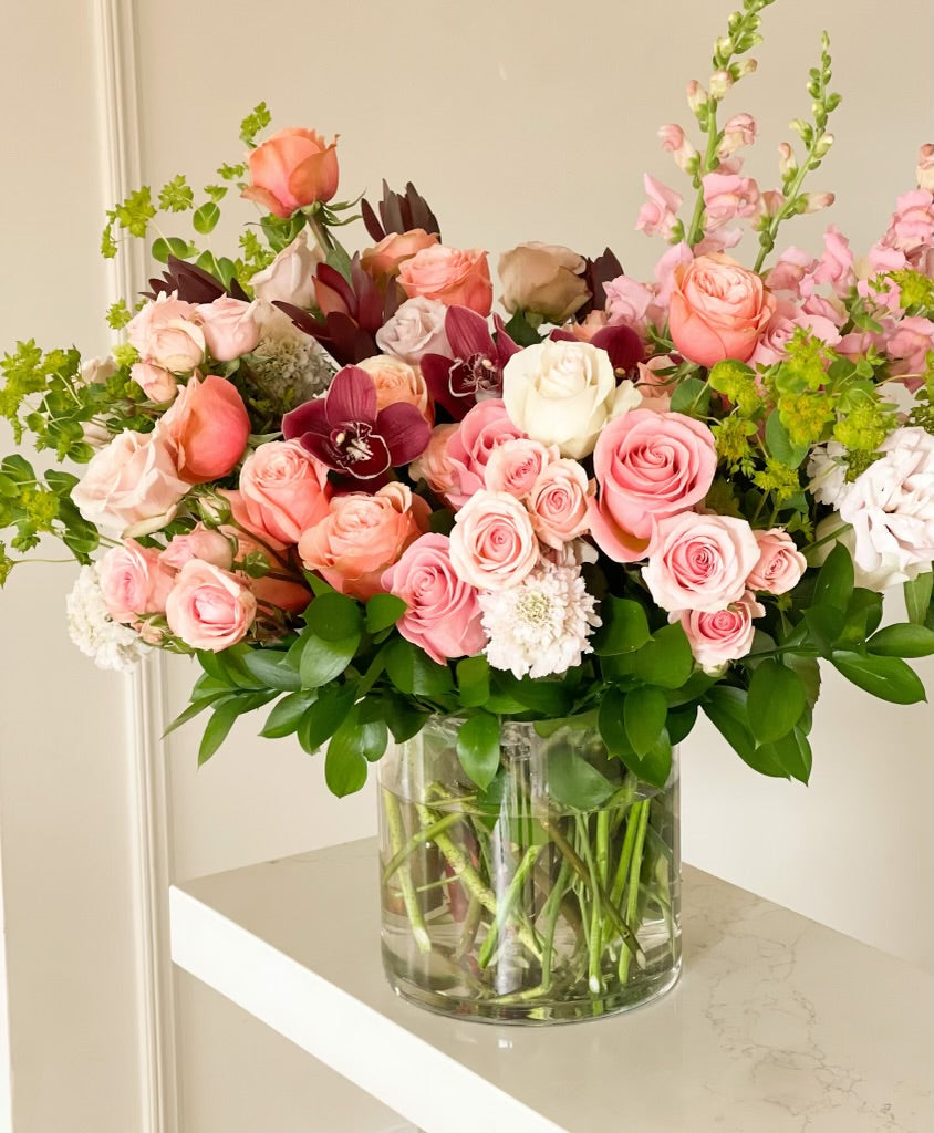 A large glass vase containing an arrangement of fresh roses, snapdragons, cymbidium orchids, and foliage in a mix of pink, red, and white colors.