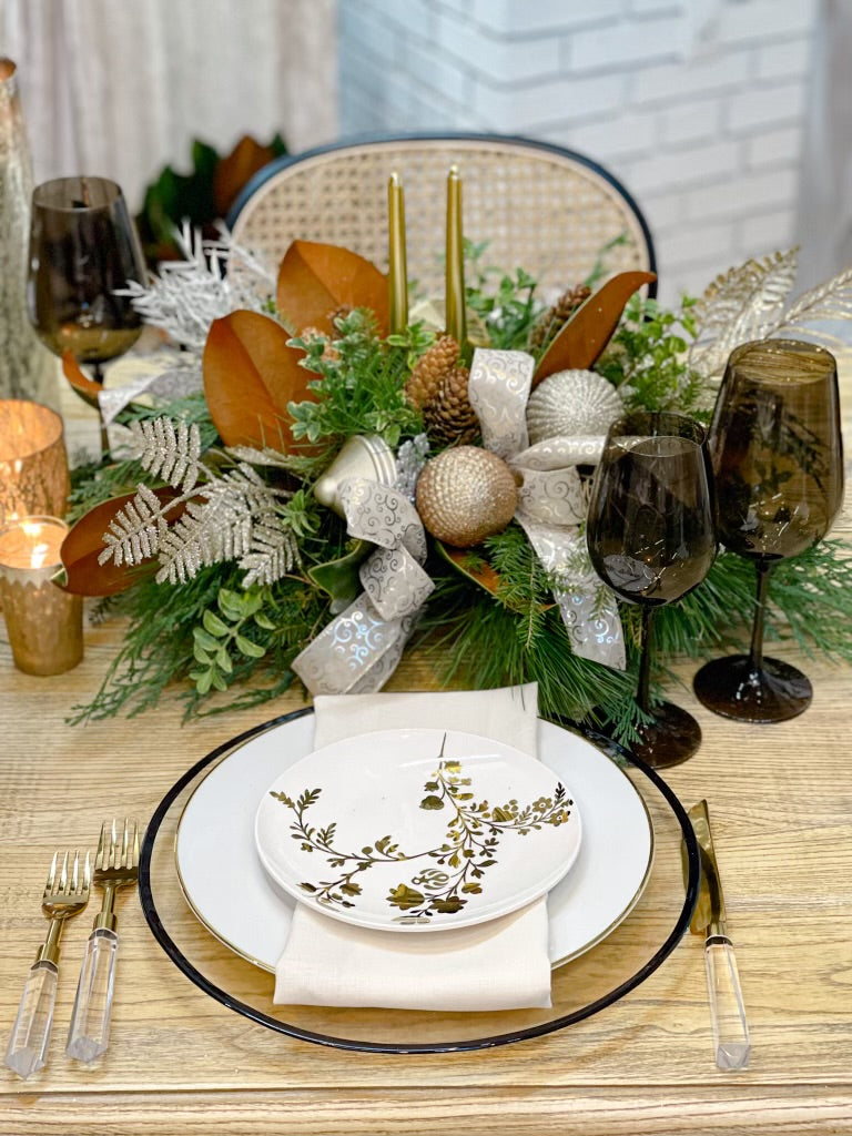 A decorative centrepiece featuring gold dipped candle sticks, pine cones, and festive ribbon, wrapped in fragrant winter greens, displayed on a table with cutlery and a plate.