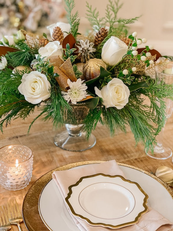 A festive centerpiece featuring white roses, berries, and greenery in a metallic glass vase, set on a table with dinnerware and candles.