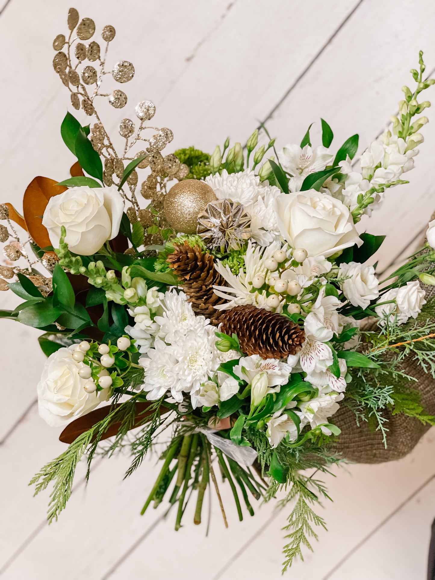 a winter flower bouquet with white roses, winter greens, pinecones, and ornaments held against a white backdrop.