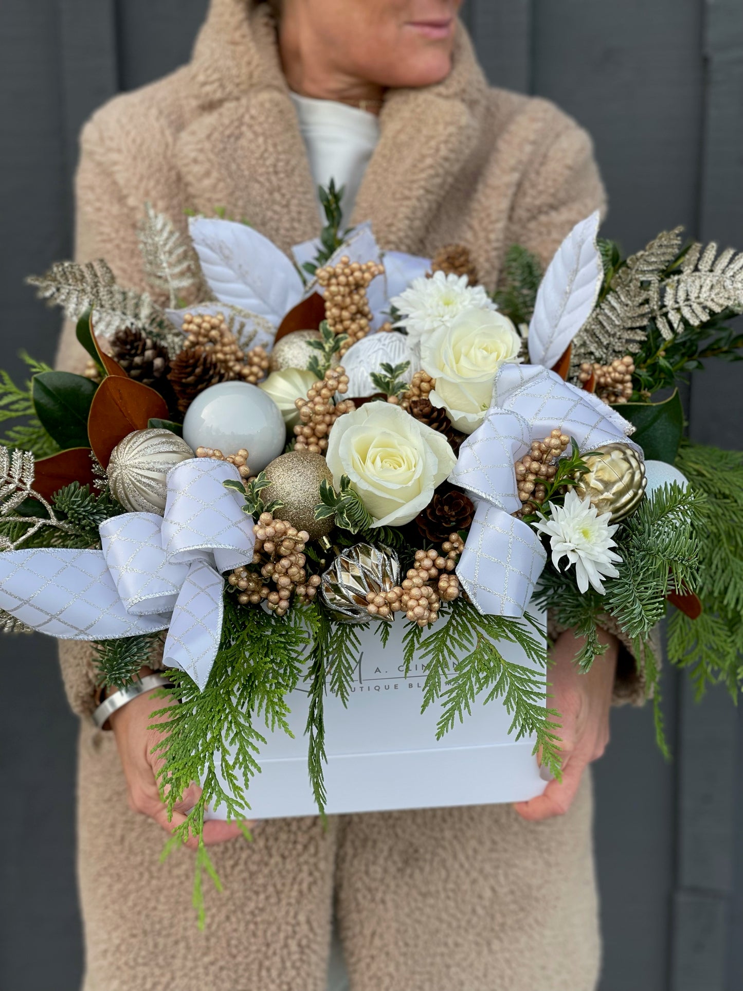 A decorative wreath held by a person, featuring a variety of green and white flowers, gold accents, and natural pinecones.