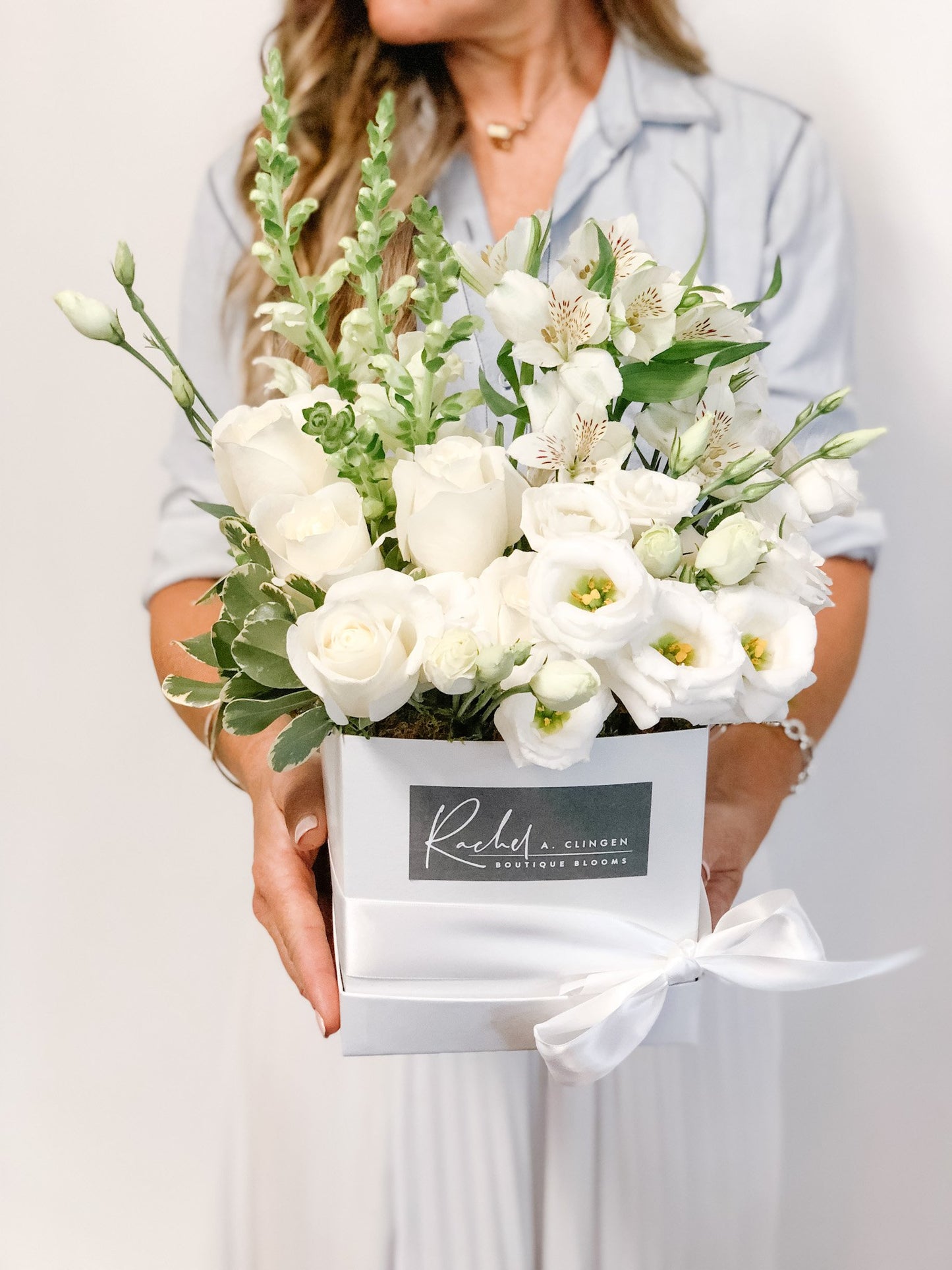 A collection of white flowers including roses and other complementary flowers, arranged in a small square pink bloom box.