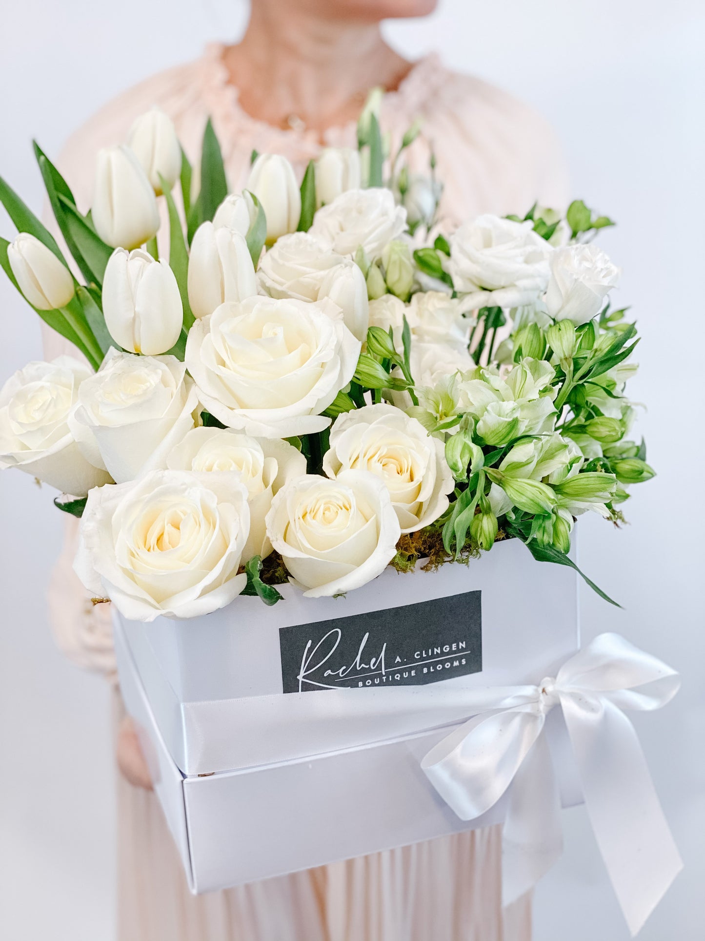 A person holding a large white floral arrangement in a rectangular box with a ribbon, containing white roses, tulips, and other premium flowers.