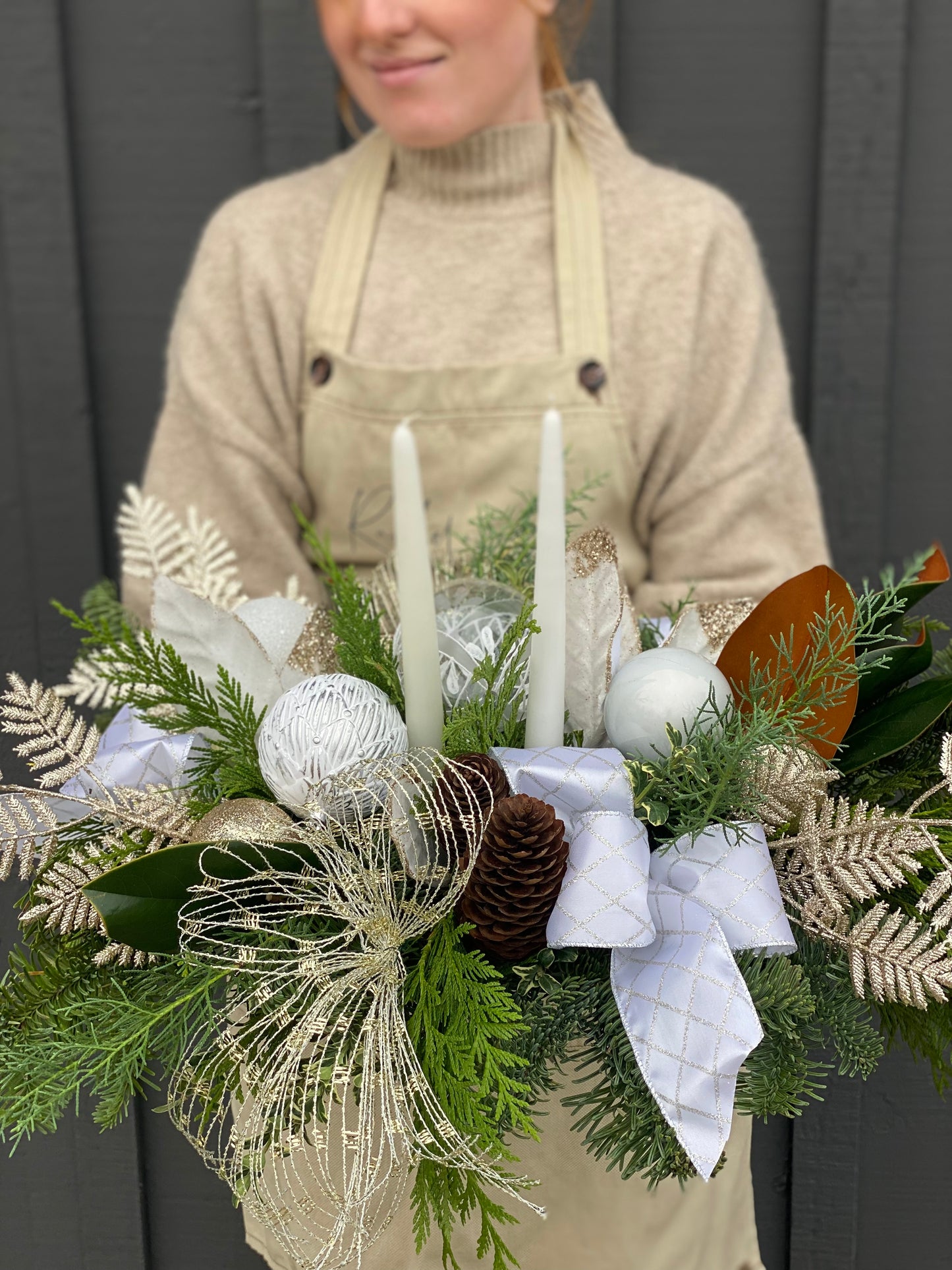 A floral arrangement featuring green and white foliage, brown pine cones, white and green ornaments, and two taper style candles, held by a person.